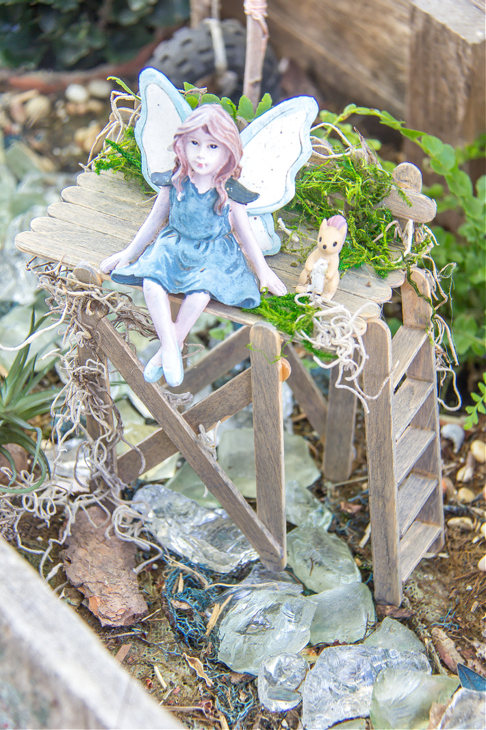 A fairy sitting on a popsicle stick viewing platform in a fairy garden.