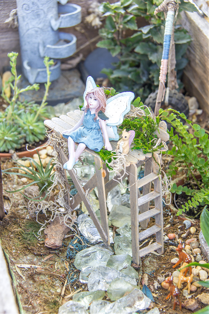 A fairy garden with a handmade popsicle stick viewing platform and a fairy sitting on it with a squirrel.