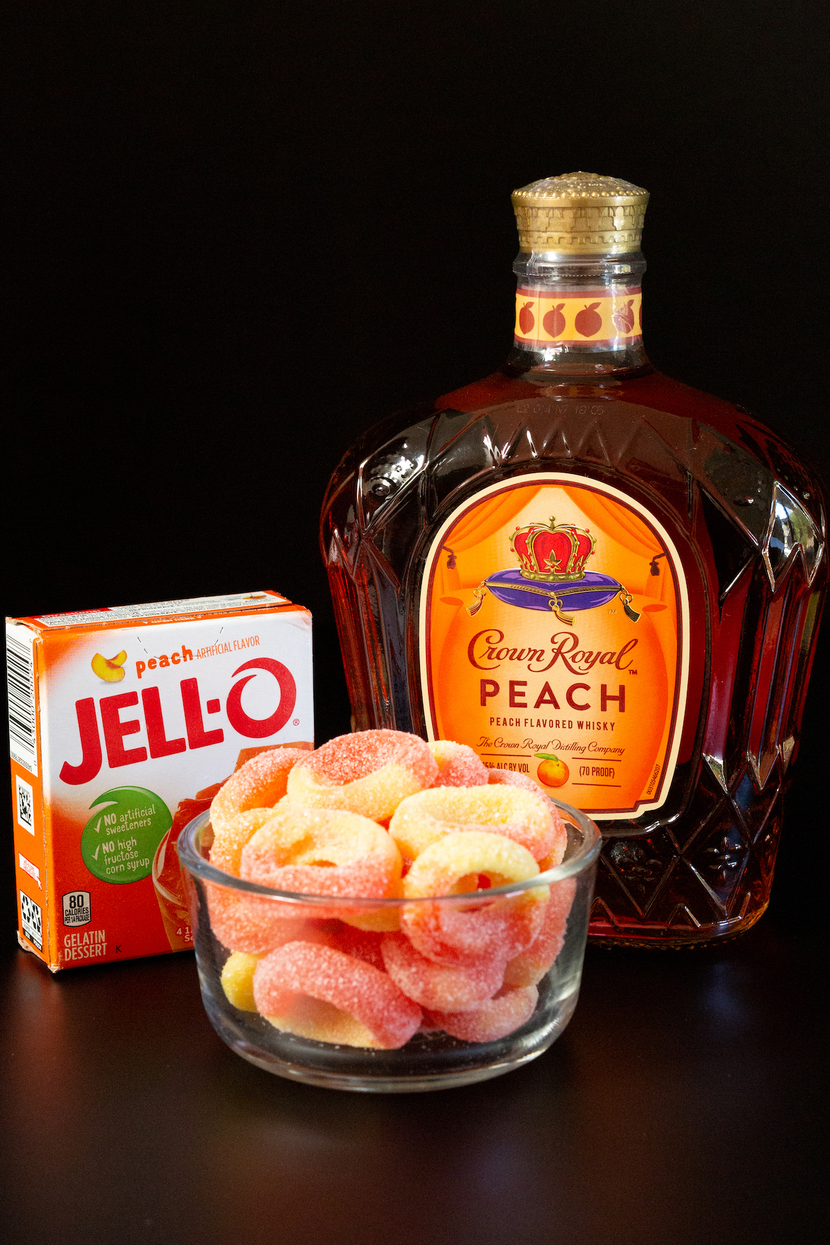 A box of peach gelatin, peach gummy rings, and a bottle of Crown Peach on a black background.