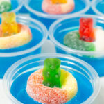 9 2oz plastic containers filled with bright blue jello topped with a candy gummy bear and gummy ring.