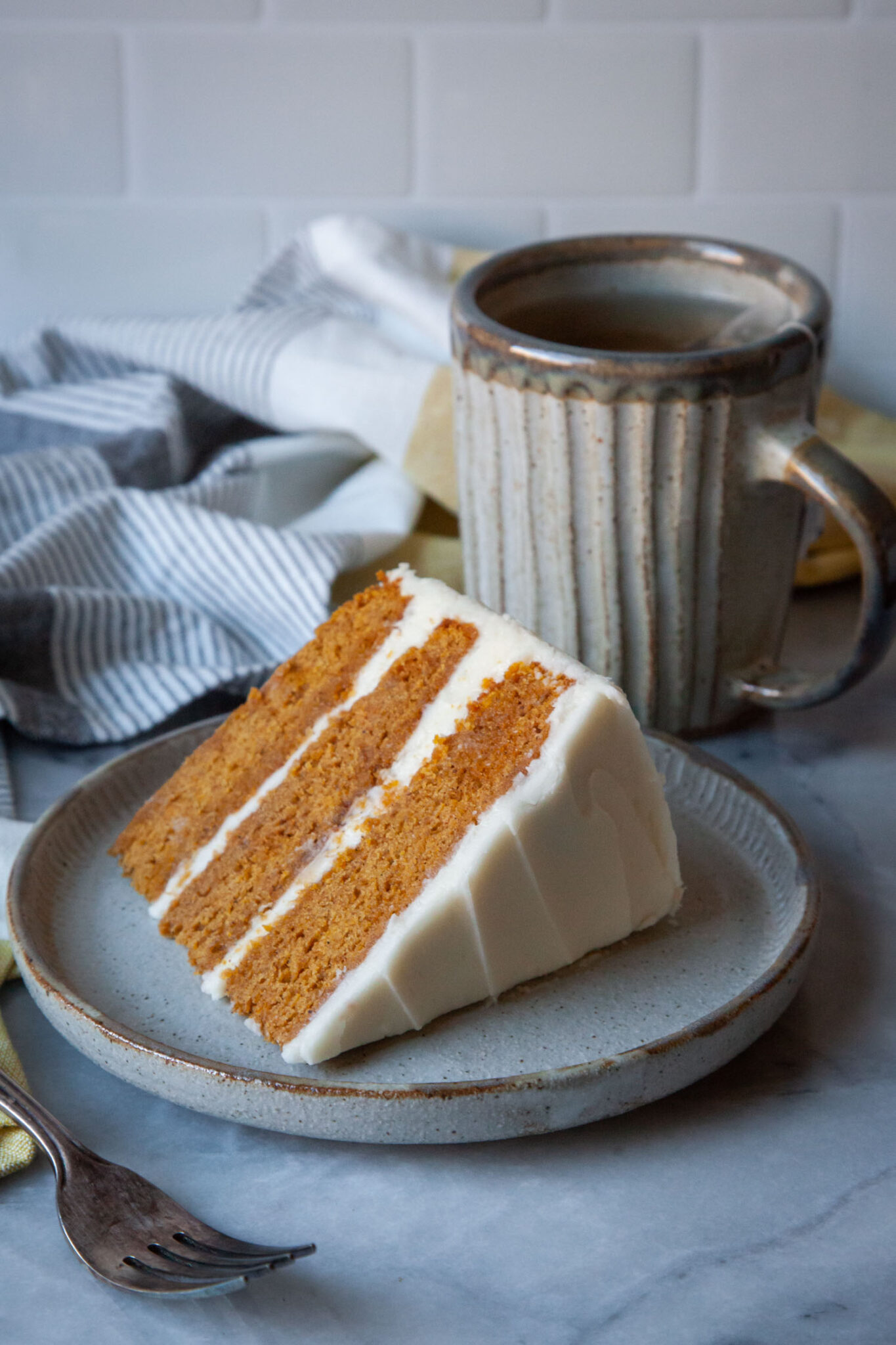 A slice of pumpkin layer cake on a plate, with a mug of tea behind it.