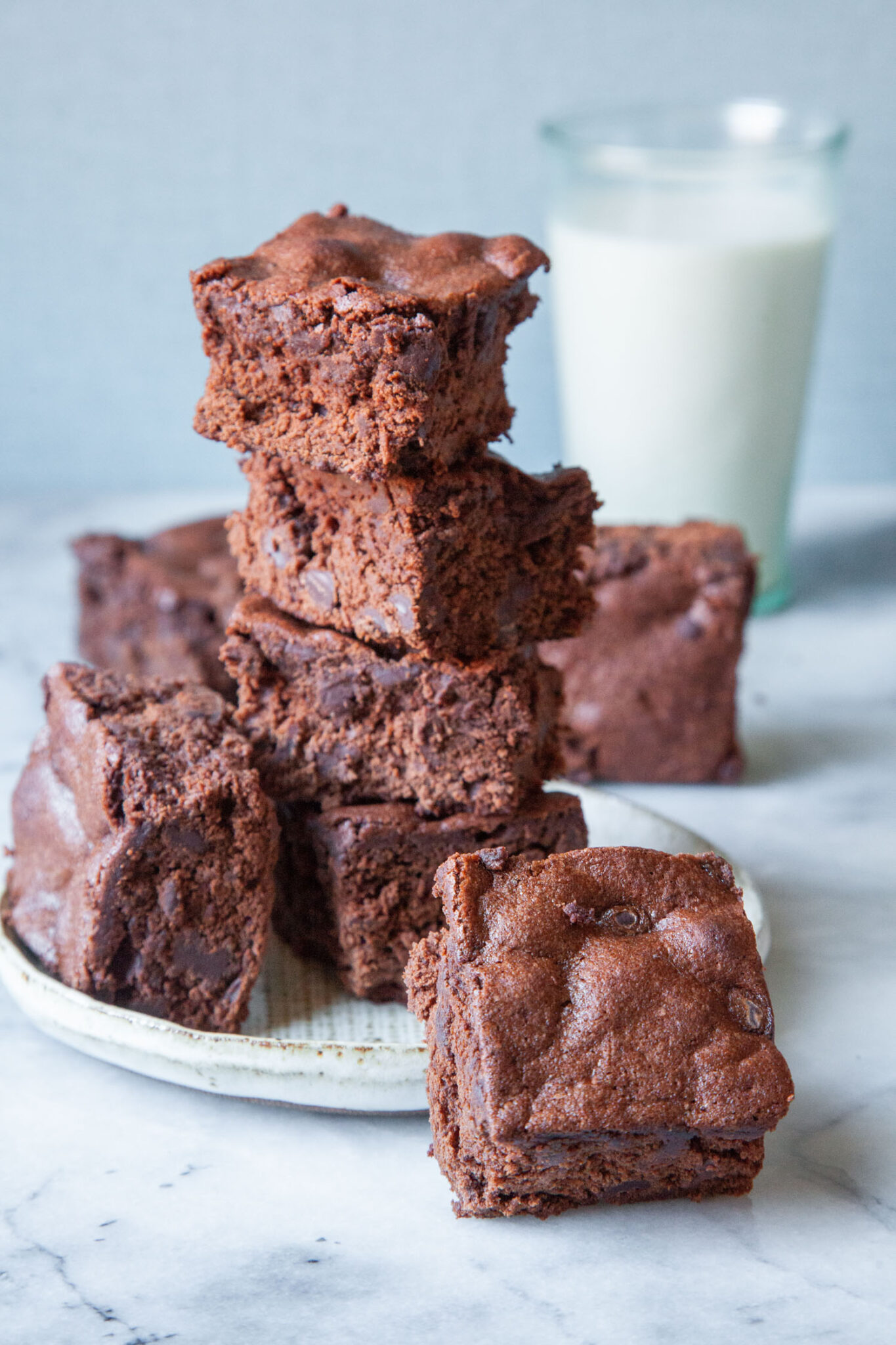 A stack of cakey brownies on a plate with one brownie tilted and sitting off the plate on the table. A glass of milk is behind the stack of brownies.