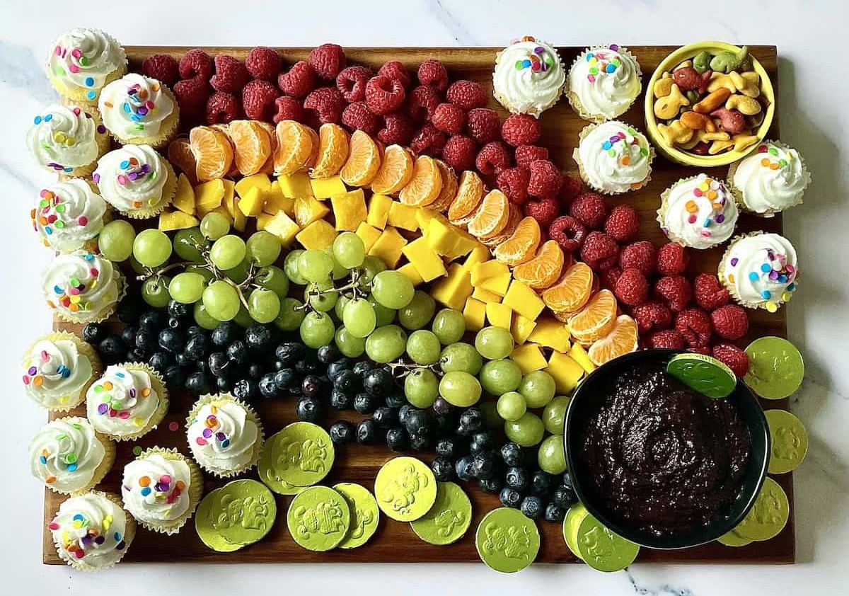 a rainbow themed charcuterie board with cupcakes, candy, fruit, and a pot of chocolate dip from tasty oven.