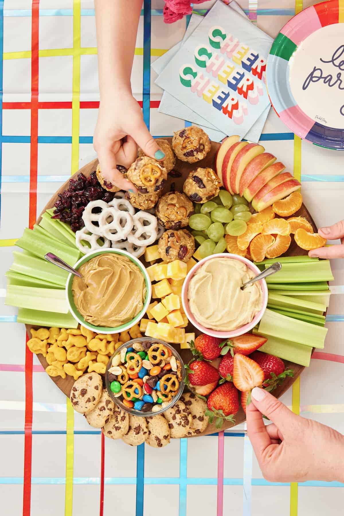 A circular wooden board with hummus, peanut butter, snack mix (made with peanuts, mini pretzels, chocolate chips and M&Ms), cheese cubes, celery chunks, apple slices, tangerine segments, green grape halves, strawberry halves, goldfish crackers, dried cranberries, yogurt-covered pretzels and Mini Chocolate Chip Cookies.