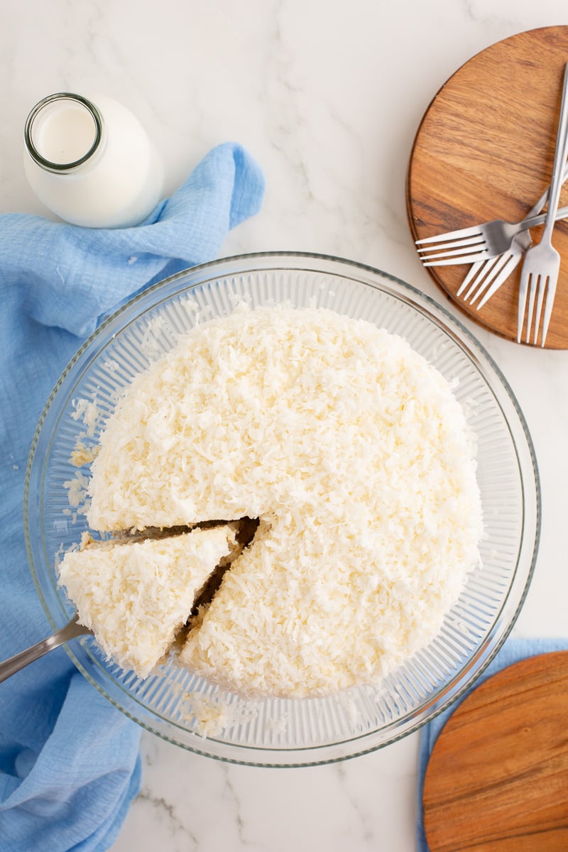 Overhead image of coconut cake on a glass cake stand with one piece being removed.